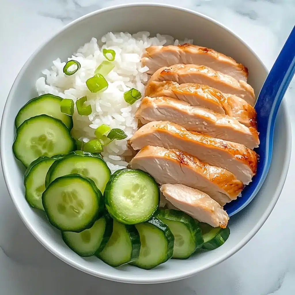 One Pot Hainanese Chicken and Rice, Hainanese Chicken and Rice, One Pot Chicken and Rice, Easy Hainanese Chicken, Fragrant Chicken and Rice A white plate holds a bed of white rice that covers the bottom layer, topped with five slices of cooked chicken laid neatly in the middle, each slice showing a light golden brown edge and smooth texture. To the right side, there is a row of thin, round cucumber slices with bright green skin and pale green centers. Scattered on top of the chicken are small pieces of chopped green onion. A dark blue spoon rests on the edge of the plate, partly on the rice, with a shiny surface reflecting light. The plate is set on a white marbled surface.
