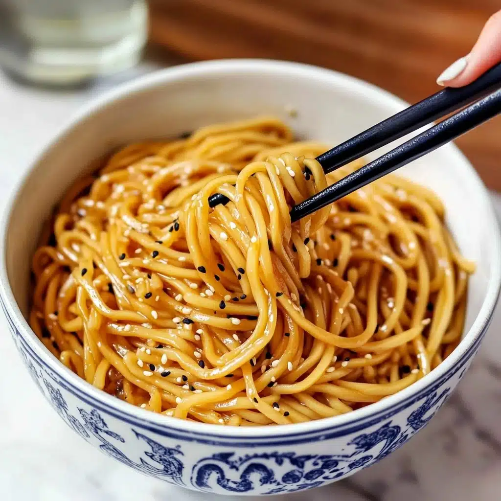 Hibachi Noodles, Hibachi Noodles recipe, easy hibachi noodles, homemade hibachi noodles, quick hibachi noodles A white bowl with blue patterns is full of thick, shiny brown noodles mixed with black and white sesame seeds. Two wooden chopsticks with black grips hold up a cluster of the noodles in the bowl. The bowl sits on a wooden table with a white cloth with blue stripes nearby and blurred bottles in the background. The surface below everything is a white marbled texture.