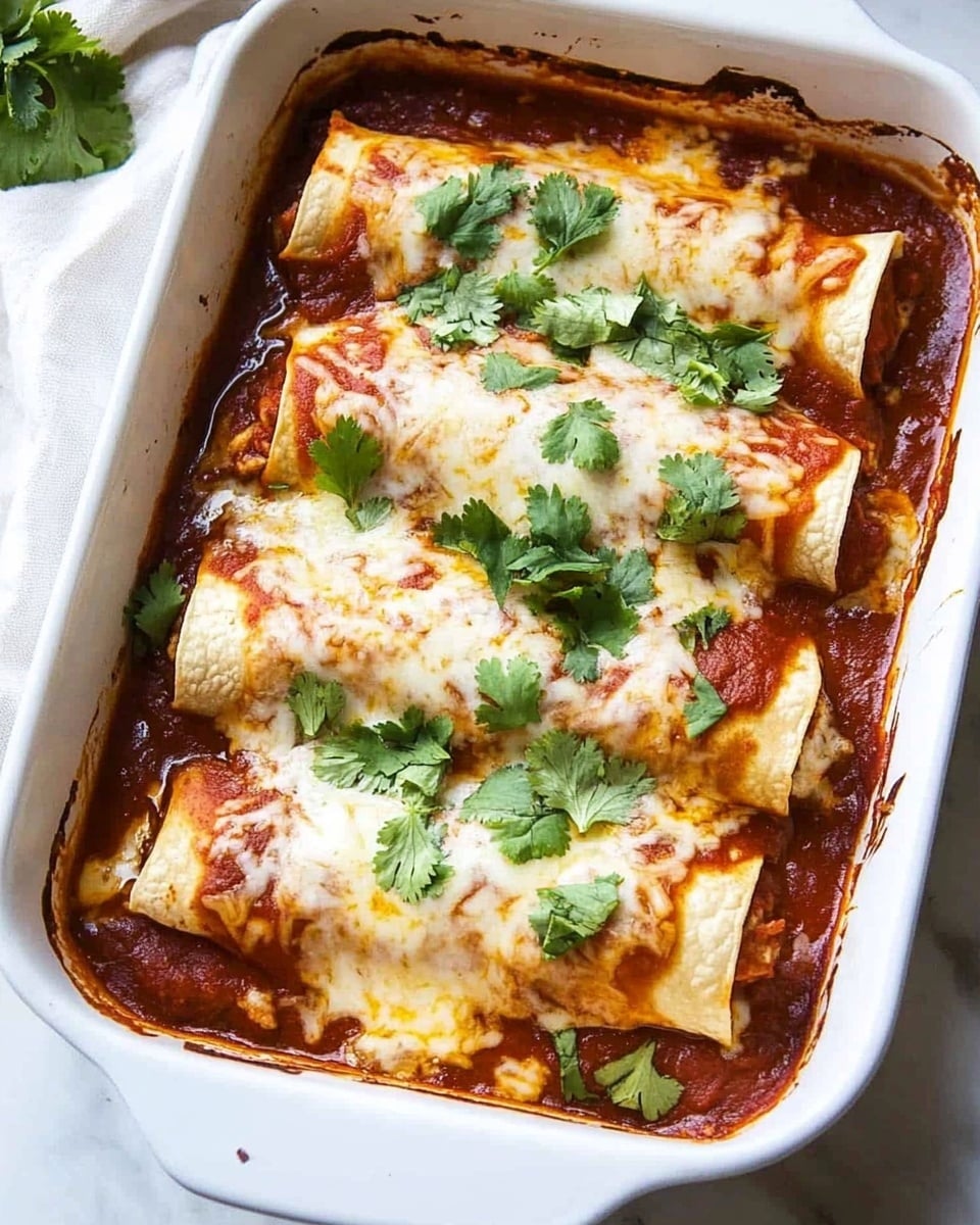Beef Enchiladas, homemade enchiladas recipe, Tex-Mex beef enchiladas, easy beef enchiladas, hearty enchilada casserole The image shows a white rectangular baking dish filled with six rolled tortillas arranged side by side in two rows of three. Each tortilla is covered with a rich red sauce that pools at the edges of the dish. On top of the tortillas is a layer of melted white cheese, slightly browned in some spots, creating a smooth, creamy texture. Fresh green cilantro leaves with uneven, natural shapes are scattered over the melted cheese, adding a fresh color contrast. The dish sits on a white marbled surface with a white cloth partially visible on the left side.