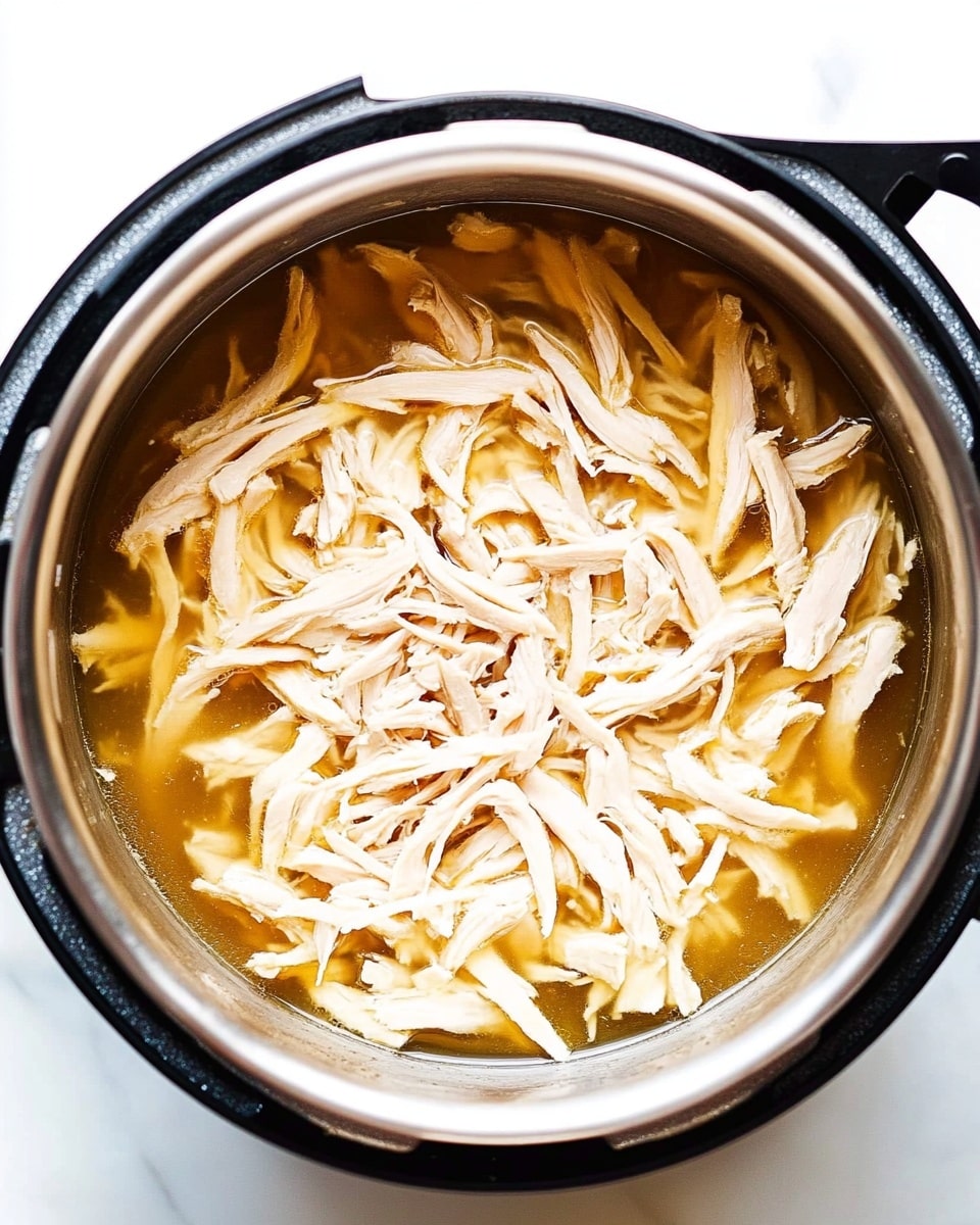 Inside a black Instant Pot with a shiny silver interior, there is one layer of shredded light beige chicken sitting in a thin layer of golden-brown broth that fills the bottom of the pot. The shredded chicken pieces are spread evenly, showing some moisture with a slight shine. The pot is placed on a white marbled surface. Photo taken with an iphone --ar 4:5 --v 6.1