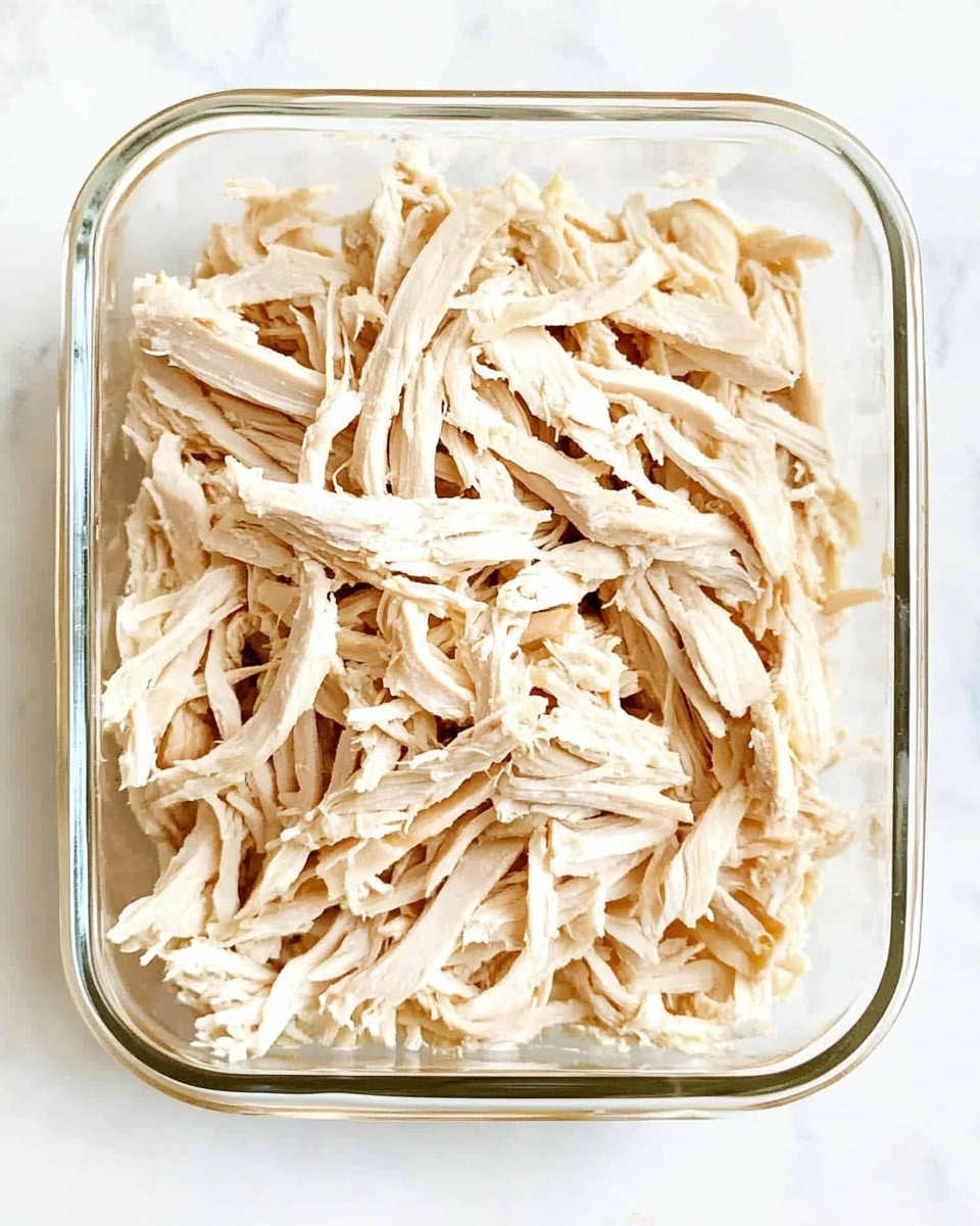 The image shows one clear glass rectangular container filled with a single layer of shredded pale beige chicken pieces. The chicken strands are uneven in size and look tender and moist. The container is placed on a white marbled surface, and no other items or layers are visible. The photo taken with an iphone --ar 4:5 --v 6.1