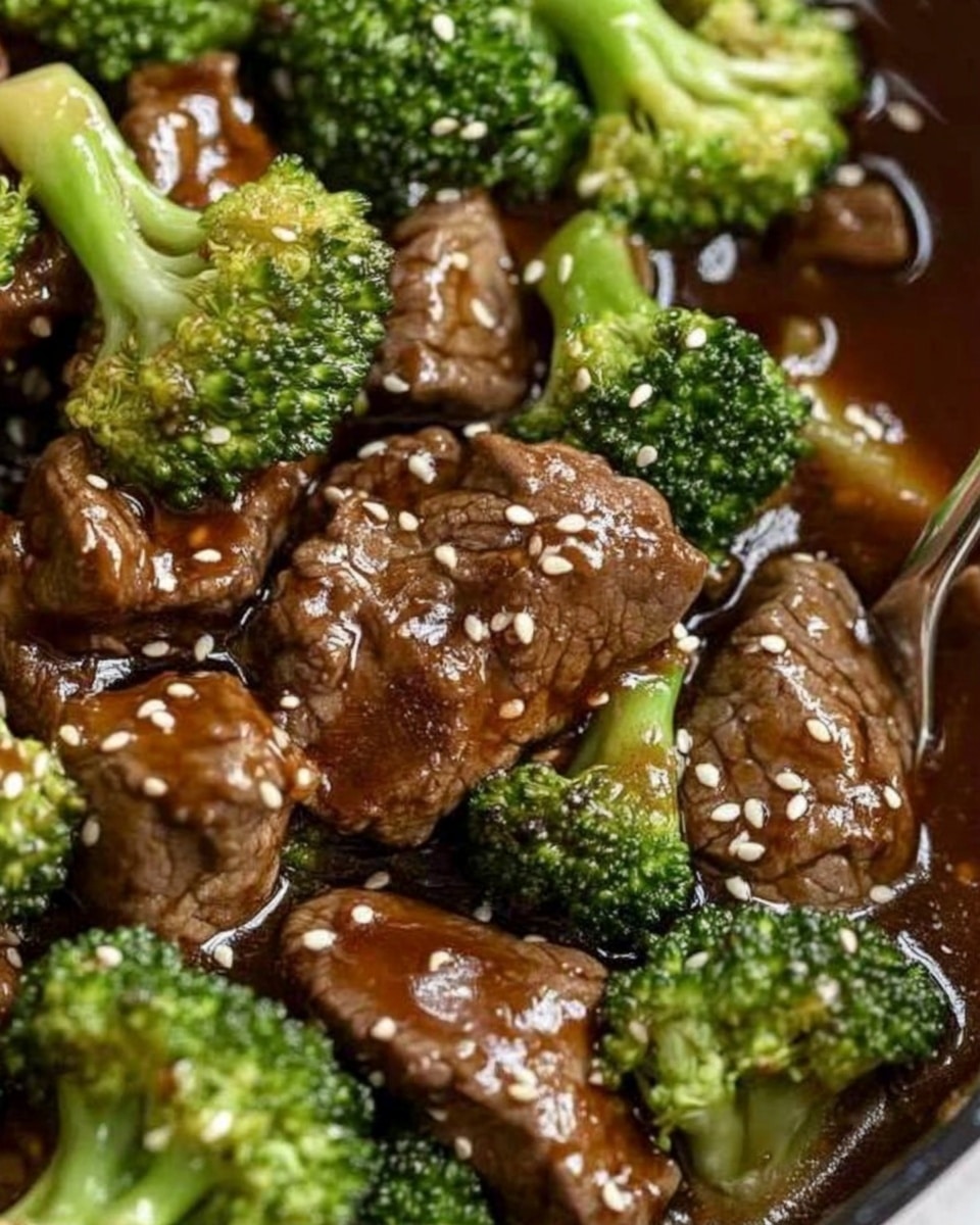 Chinese Beef and Broccoli Stir Fry Asian stir fry healthy quick dinner savory beef and broccoli A close-up of a white plate filled with a layer of white rice at the bottom, topped with pieces of cooked beef and bright green broccoli florets. The beef and broccoli are coated with a thick, shiny brown sauce that pools slightly on the rice. White sesame seeds are sprinkled over the top, adding small contrasting dots. The plate sits on a white marbled textured surface, with a pair of wooden chopsticks resting on the plate's edge.