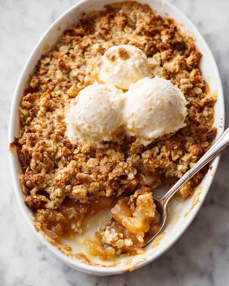The image shows a white oval dish filled with a warm fruit crumble dessert. The bottom layer is soft, golden fruit pieces that look juicy and tender. On top is a thick, crunchy oat topping with a golden-brown color and a crumbly texture, some parts are slightly darker from baking. Two melting scoops of creamy white ice cream sit on the surface, softening and blending slightly into the crumble. A spoon rests inside the dish, scooping up a mix of the fruit and oat layers, showing the moist texture underneath. The dish is placed on a white marbled surface. Photo taken with an iphone --ar 4:5 --v 7