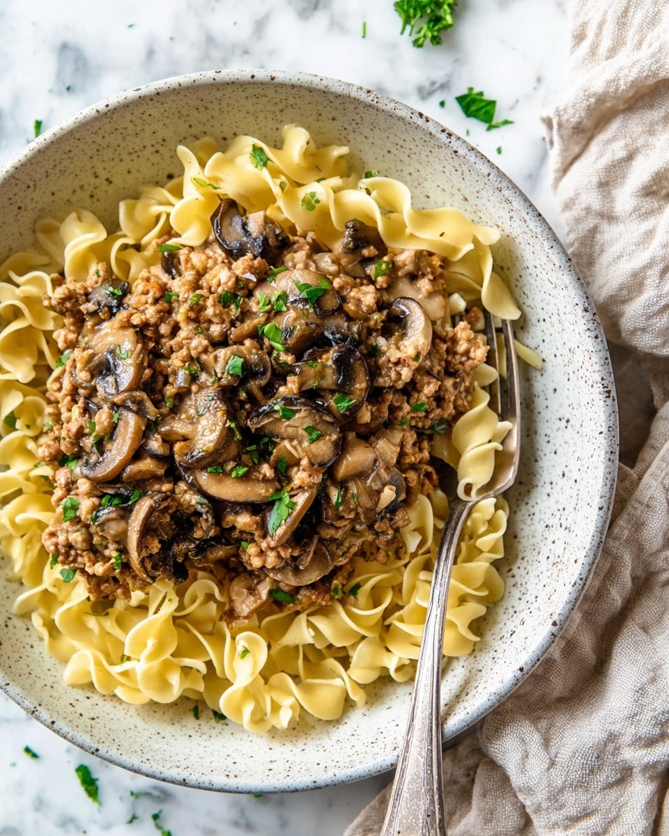 A white speckled bowl filled with three layers: the bottom layer has yellow twisted egg noodles, the middle layer has browned minced meat mixed with golden cooked onions, and the top layer has dark brown sliced mushrooms scattered all over, sprinkled with chopped green parsley. A silver fork rests on the right side, partially in the bowl, with some noodles wrapped around it. The bowl sits on a white marbled surface with a beige cloth napkin in the top right corner. Photo taken with an iphone --ar 4:5 --v 7
