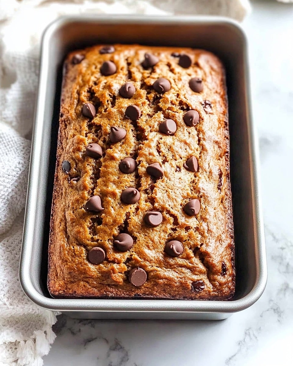 A freshly baked loaf of banana bread sits in a metal rectangular baking pan lined with parchment paper on one side, its top golden brown with a slightly rough texture. The bread is thick with a cracked surface, generously sprinkled with dark chocolate chips that are slightly melted and scattered evenly across the top layer. The pan is placed on a white marbled surface with a soft white cloth partially visible in the top left corner. Photo taken with an iphone --ar 4:5 --v 6.1