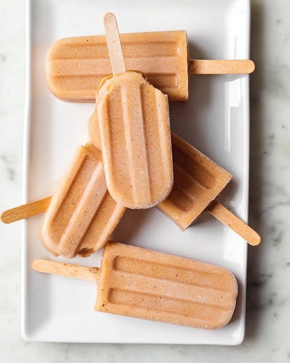 Five light brown popsicles with wooden sticks are placed on a white rectangular plate. One popsicle is held by a woman's hand and shows a bite taken from the top, revealing a slightly darker creamy inside. The popsicles have a smooth, slightly frosty texture. The plate is on a white marbled surface. photo taken with an iphone --ar 4:5 --v 7