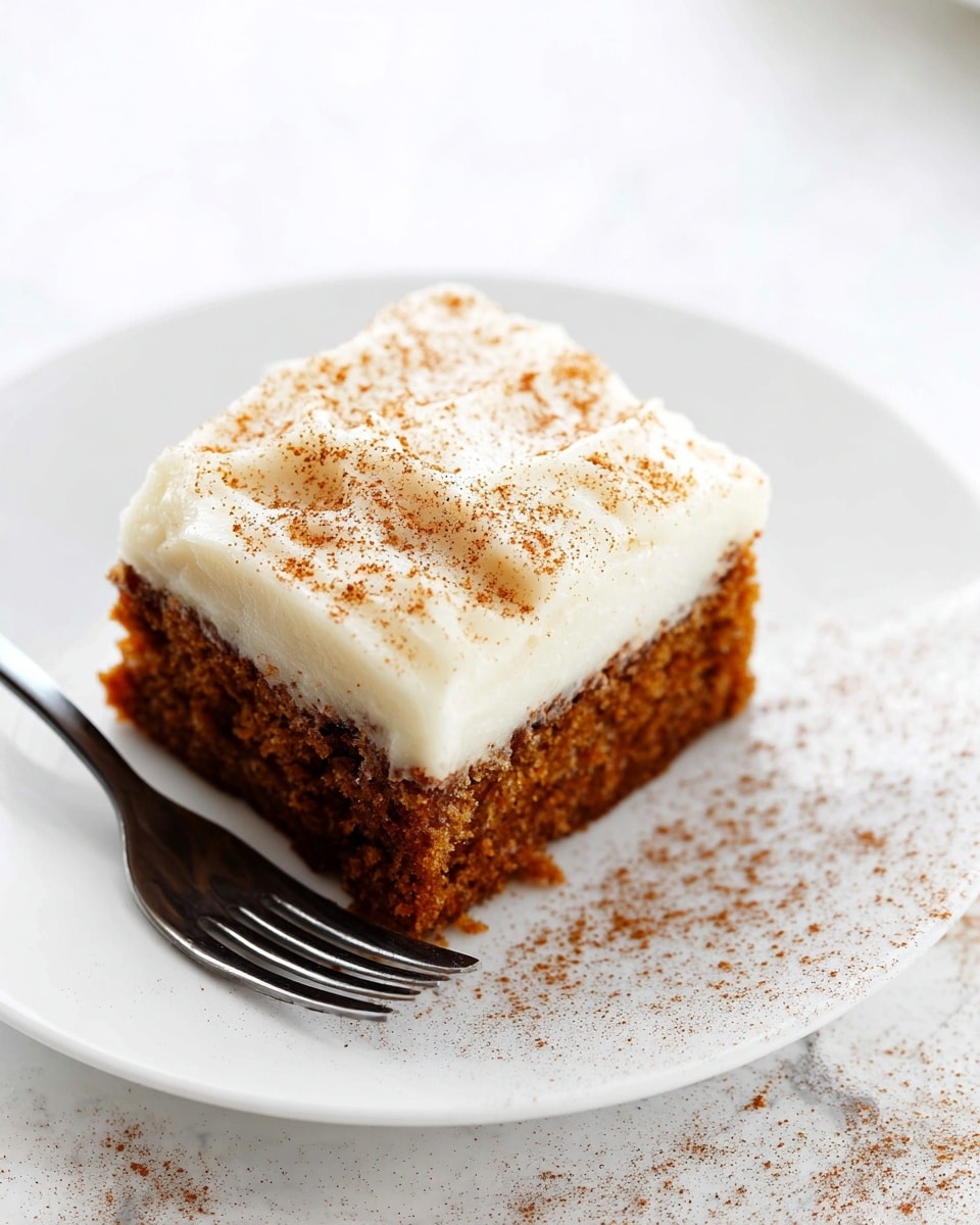 A square piece of carrot cake with two layers sits on a white plate. The bottom layer is moist and dark orange-brown with a rough texture showing bits of carrot. On top is a thick, creamy white frosting layer, smooth with light brown cinnamon powder sprinkled over it. Next to the cake is a silver fork resting on the plate. The scene is set on a white marbled surface with soft natural light and another plate blurred in the back. Photo taken with an iphone --ar 4:5 --v 7