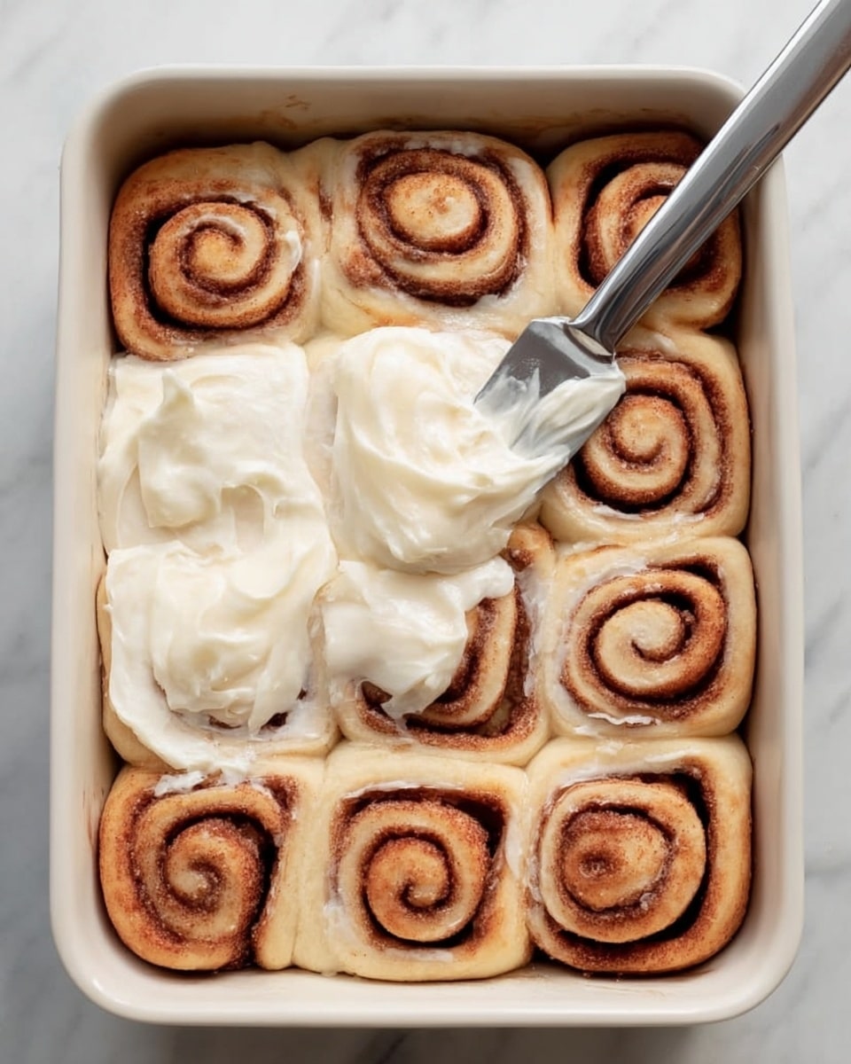 A white rectangular baking dish holds twelve cinnamon rolls arranged in a 3 by 4 grid. Each roll shows distinct dark brown cinnamon spirals on a pale dough base. In the center, a thick layer of creamy white frosting is being spread with a metal knife, held by a woman's hand at the bottom right corner. The dish rests on a white marbled surface with a folded blue cloth to the left. Photo taken with an iphone --ar 4:5 --v 7