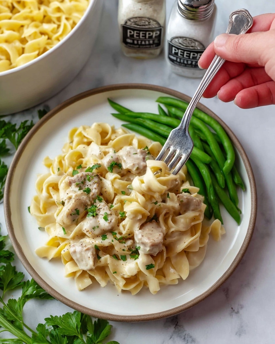 A white plate holds three main layers: on one side, light yellow egg noodles with a soft, slightly curled texture mixed with pieces of tender white chicken, lightly sprinkled with small green herbs and black pepper; next to the noodles is a bright green layer of steamed green beans cut into bite-sized pieces with a smooth and shiny surface; a woman's hand is using a silver fork to lift a small bundle of noodles and chicken from the middle of the plate. The plate sits on a white marbled surface with some green parsley leaves nearby, and in the background, salt and pepper shakers and a pot of more noodles are partially visible. Photo taken with an iphone --ar 4:5 --v 7