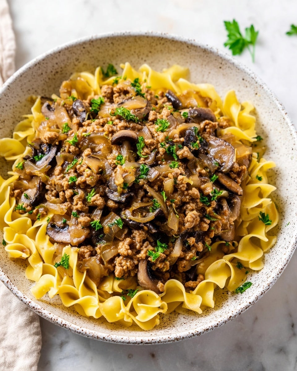 A white speckled bowl holds a dish with two main layers; the bottom layer is yellow egg noodles with a soft, curly texture filling the bowl, while the top layer is a thick ground turkey sauce mixed with sautéed dark brown mushrooms and translucent cooked onions, sprinkled with fresh chopped green parsley. The bowl sits on a white marbled surface with some scattered parsley leaves nearby, and a beige cloth with dark stripes is softly blurred in the background, photo taken with an iphone --ar 4:5 --v 7