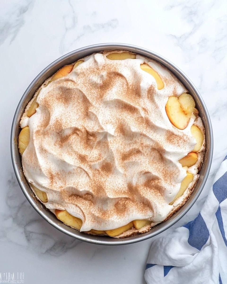 A round baking pan filled with two layers is shown from above on a white marbled surface. The bottom layer is made of chunky golden yellow fruit pieces, visible around the edges. The top layer is a thick, light beige cream covered with a dusting of fine brown cinnamon powder, spread unevenly to show small swirls and texture on its surface. A white and blue checkered cloth lies partially visible on the bottom right side. photo taken with an iphone --ar 4:5 --v 6.1