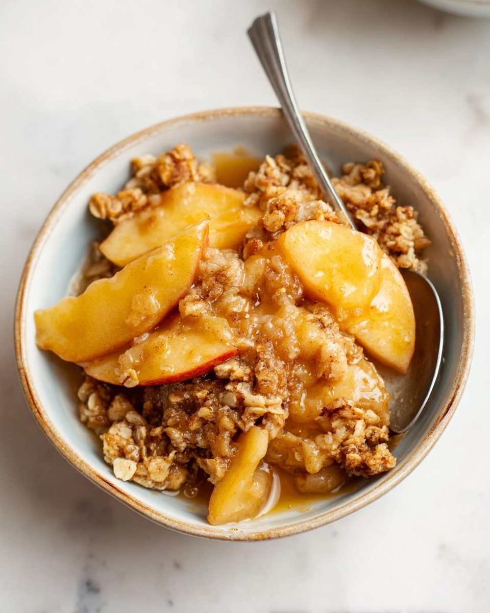 A close-up view of a bowl filled with warm apple crisp. The dish has two main layers: the bottom layer is soft, golden-brown apple slices showing a slightly translucent texture, and the top layer is a crumbly oat crumble topping in light to medium brown colors with visible oats and sugar clusters, adding crunch. The bowl is white, and a silver spoon is partially placed inside it. The background shows a white marbled surface with a soft focus on other bowls and sliced apples. Photo taken with an iphone --ar 4:5 --v 7