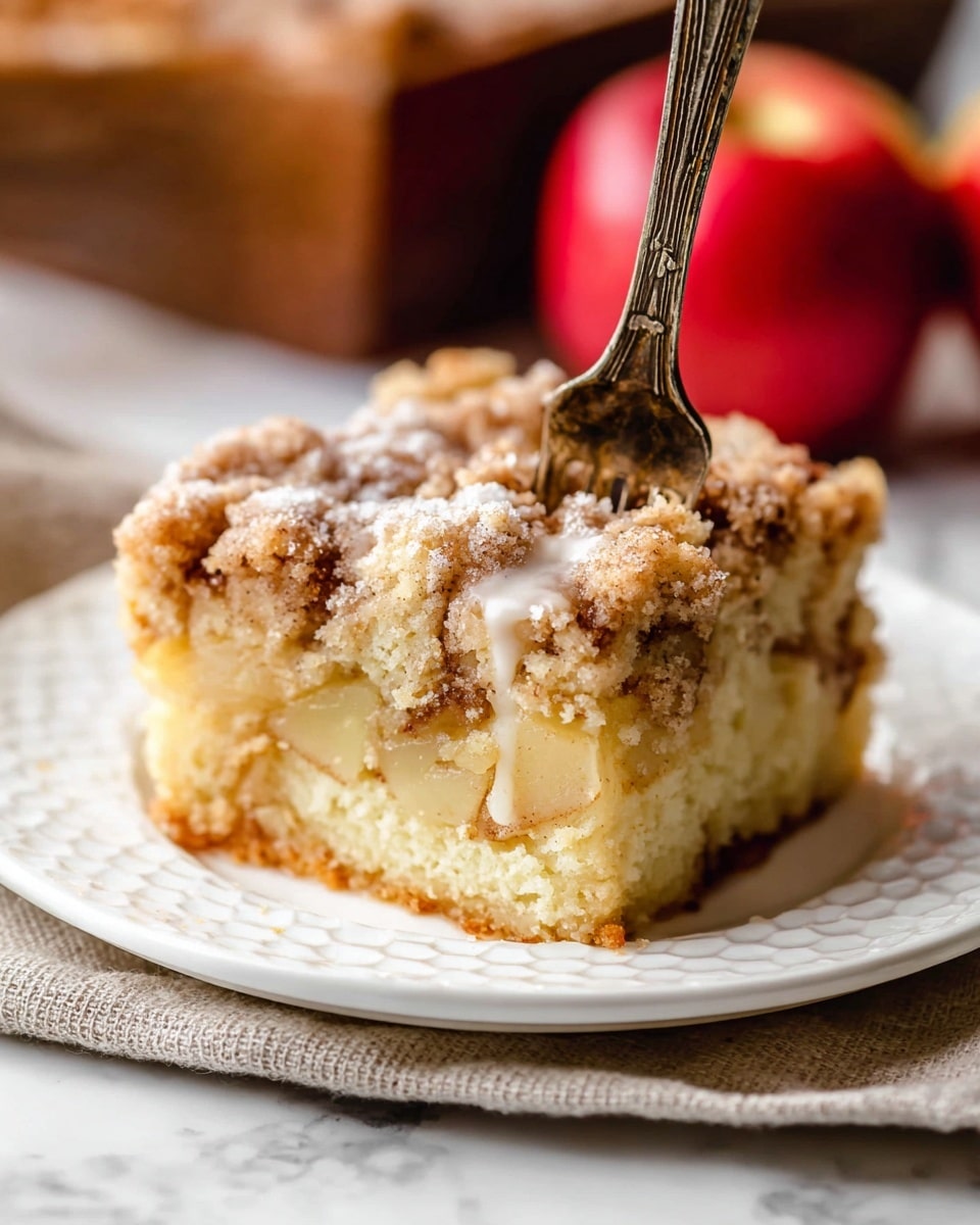 A thick square piece of crumbly apple cake sits in the center of a white plate, showing three distinct layers: a soft light yellow cake base with small chunks of apple inside, a middle layer with more apple pieces mixed into the cake texture, and a top layer of golden-brown crumb topping with white icing drizzled unevenly over it; a bronze-colored fork stands upright, pressed into the middle of the cake, and behind the plate, there are two red apples out of focus, all placed on a white marbled surface photo taken with an iphone --ar 4:5 --v 7