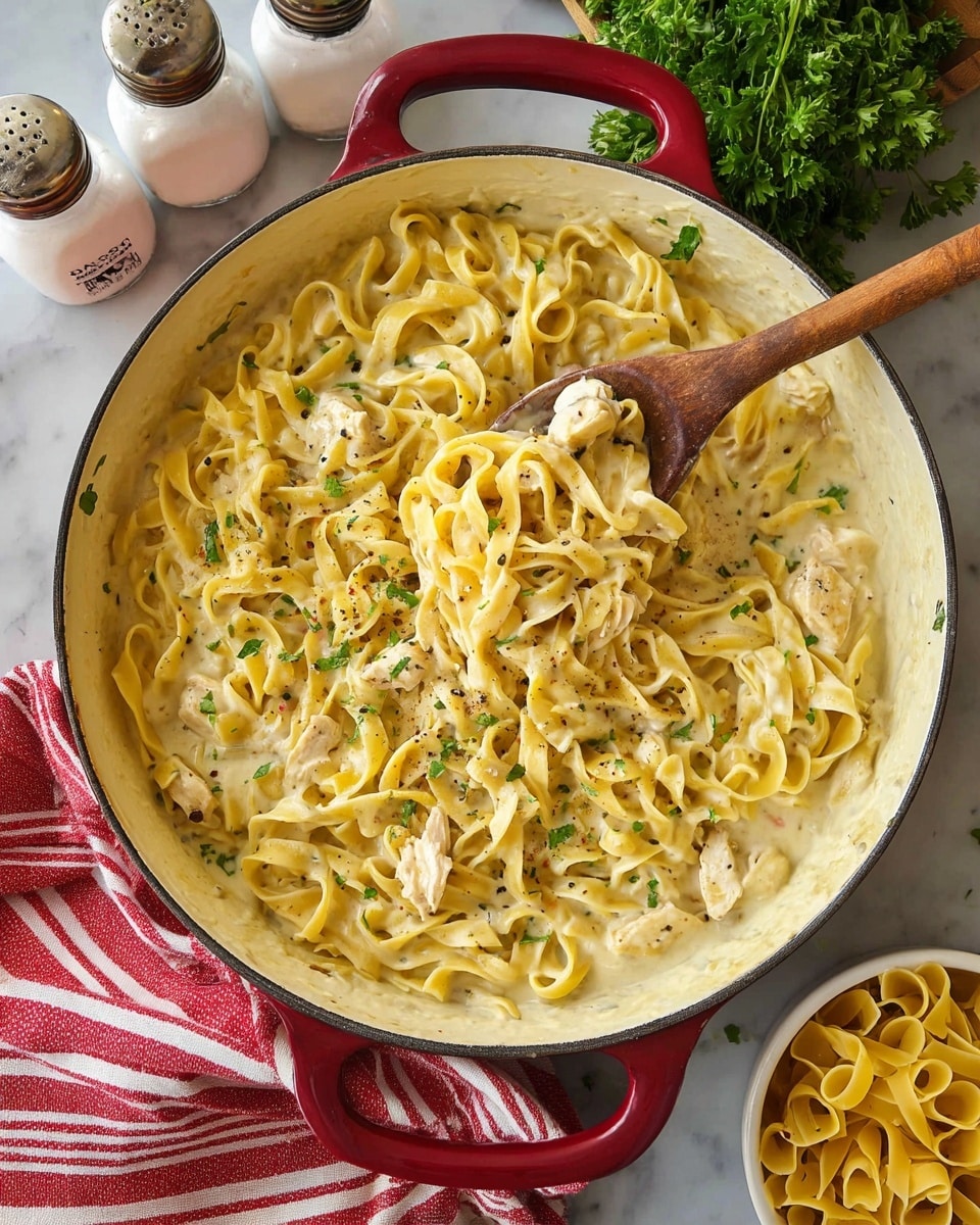 A large white pan with red handles holds creamy yellow pasta noodles mixed with small white meat pieces, likely chicken, scattered evenly throughout. The pasta is coated with a smooth, slightly shiny sauce and sprinkled with small green herbs and black pepper flakes. A wooden spoon with a wide flat head rests inside the pan, partially lifting some noodles to reveal the sauce underneath. Around the pan, there are some green parsley leaves and a red-and-white striped cloth on a dark wooden surface, with two white containers labeled salt and pepper and a small white bowl with more uncooked pasta visible in the background. Photo taken with an iphone --ar 4:5 --v 7