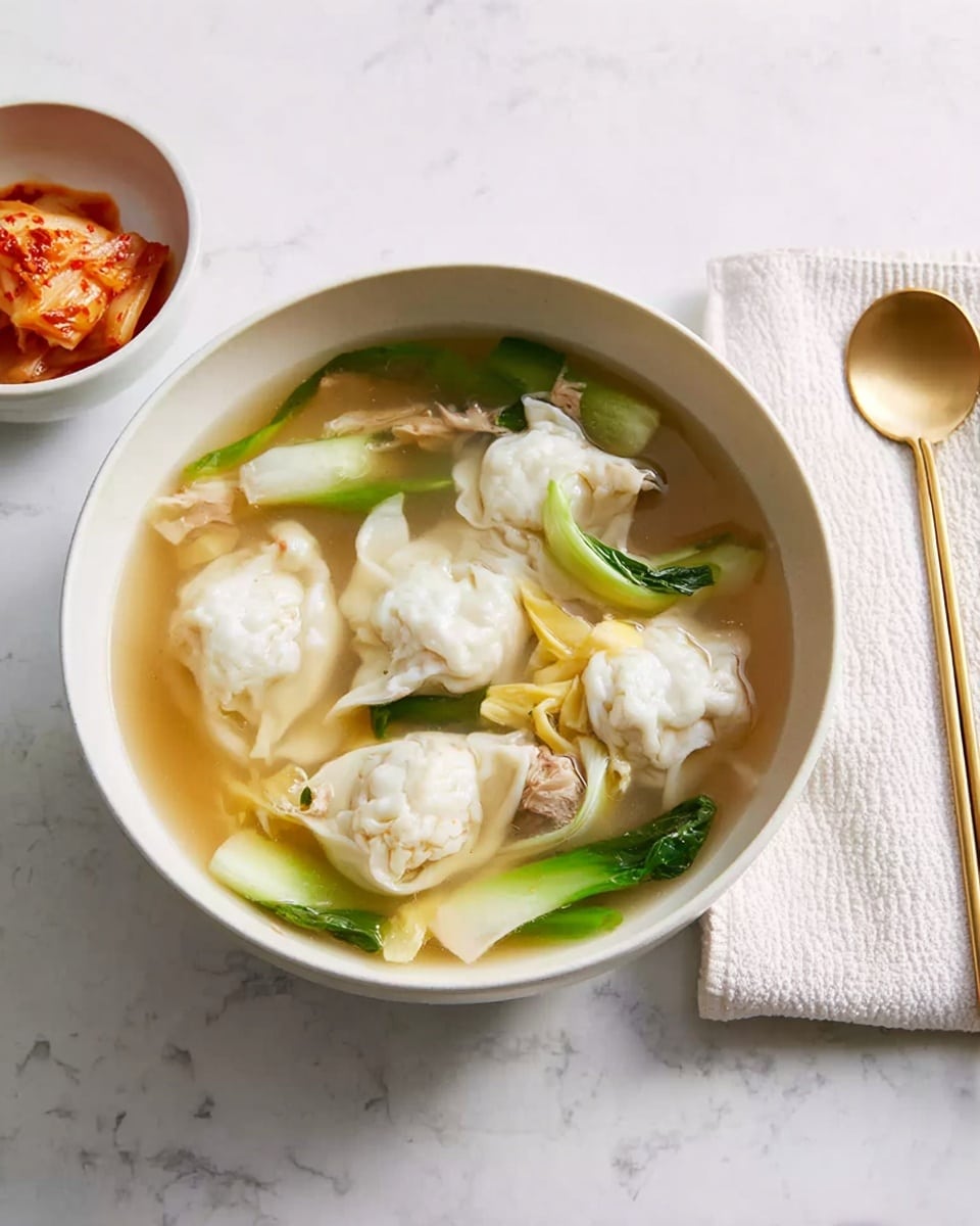 The image shows a white bowl filled with clear broth soup containing five dumplings that are white with a soft, slightly wrinkled texture. Inside the soup, there are pieces of cooked egg forming soft yellow and white ribbons, along with green onion stalks cut in halves placed around the dumplings. Some small pieces of meat are visible under the egg ribbons. To the left, a smaller white bowl holds reddish-orange kimchi with visible Napa cabbage leaves and seasoning. The bowls and utensils are set on a white marbled surface. To the right of the soup bowl, there is a white cloth napkin with a golden spoon and chopsticks resting on top. photo taken with an iphone --ar 4:5 --v 7