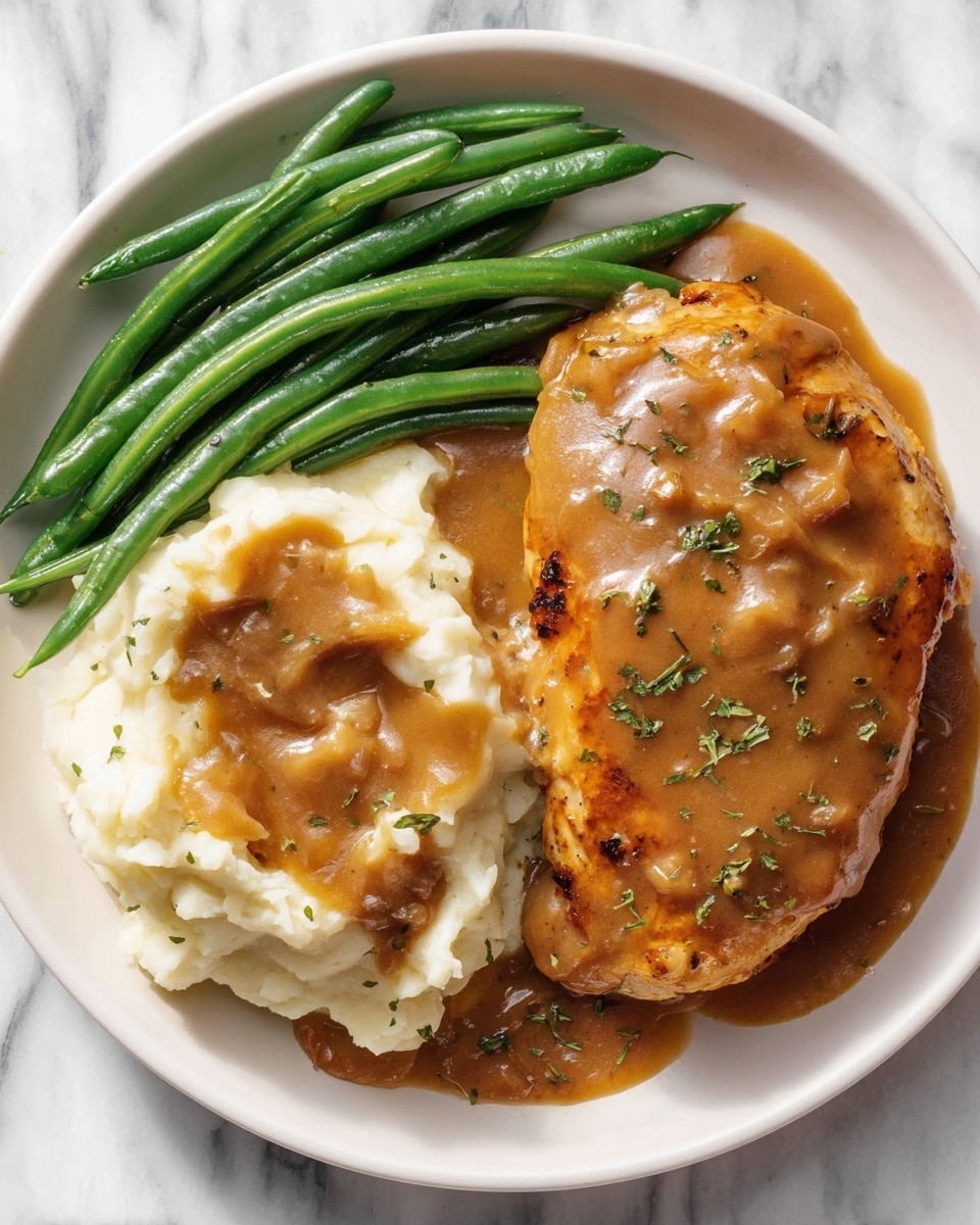 A white plate holds three parts: on the left side, bright green steamed green beans, smooth and shiny, placed close together; in the middle and right side, a large piece of golden brown cooked chicken with a thick, rich brown gravy sauce with visible bits of onion and herbs on top, partially covering creamy white mashed potatoes that have a soft, fluffy texture. The gravy sauce also covers a large portion of the mashed potatoes, and small green herb bits are sprinkled over the dish. The plate sits on a white marbled surface. Photo taken with an iphone --ar 4:5 --v 7