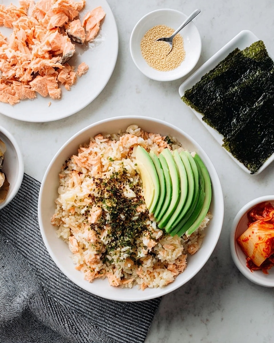 A close-up image shows a woman's hand holding a small food piece made of a dark green seaweed sheet layered with light brown cooked rice mixed with flakes of pinkish-orange salmon, a bright green avocado slice, and some reddish kimchi on top, with tiny light-colored sesame seeds sprinkled over. In the blurred background, there is a white bowl filled with the mixed rice and salmon topped with sliced avocado, a white plate with more salmon pieces, a small white dish with sesame seeds and a spoon, and a white marbled surface under everything. photo taken with an iphone --ar 4:5 --v 7