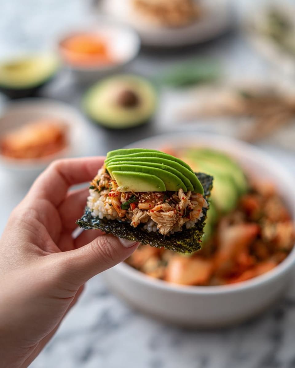 A white bowl filled with a base layer of cooked rice mixed with shredded cooked salmon, giving a light brown and white texture. On one side of the bowl, there is a layer of five slices of bright green avocado fanned out neatly. The rice mixture is sprinkled with small dark green and black seaweed flakes. Around the bowl, on a white marbled surface, there are small white plates holding extra ingredients: one with square sheets of dark green shiny seaweed, another with a small scoop of red-orange kimchi, another with salmon flakes, and a small white dish with light yellow flakes. A gray and white striped cloth is placed below on the left side. Photo taken with an iphone --ar 4:5 --v 7