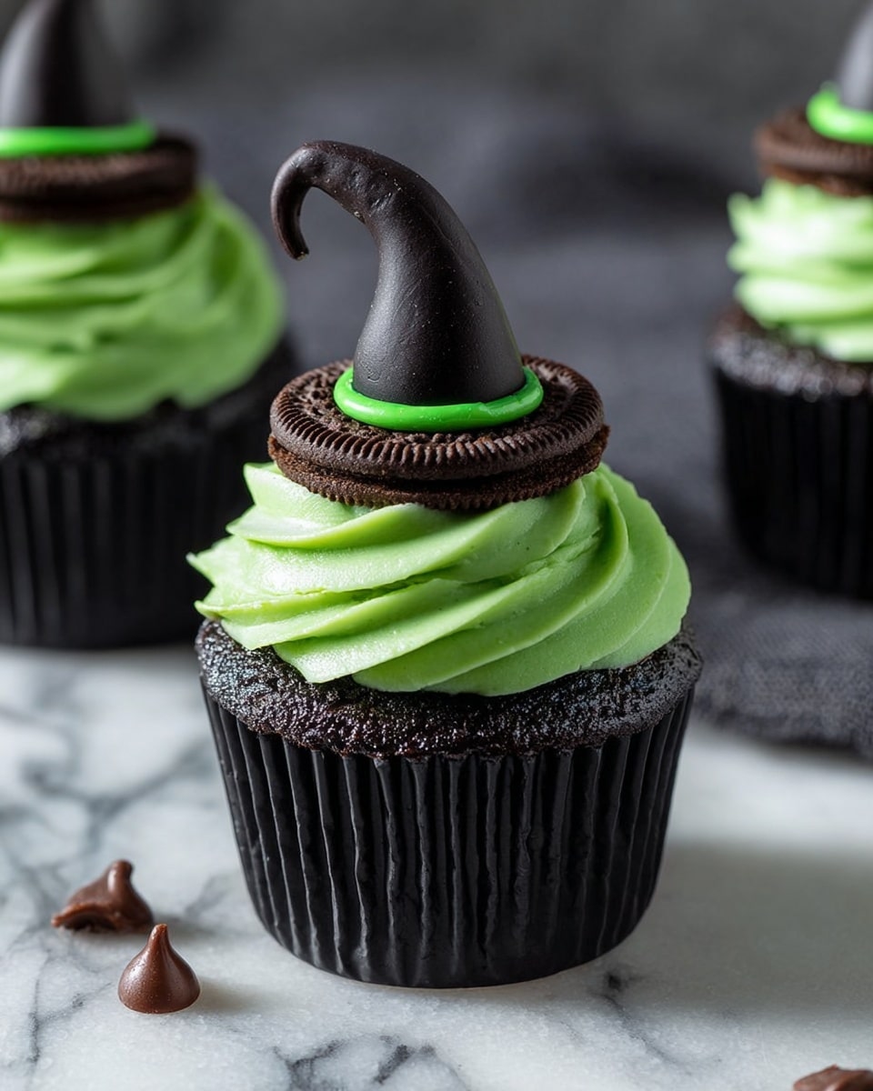 The image shows dark chocolate cupcakes with three main layers. The bottom layer is a dark chocolate cupcake in a black paper cup. On top of this is a thick swirl of bright green frosting with a smooth and creamy texture. Sitting on the frosting is a dark chocolate cookie, and on top of the cookie is a small black witch hat made of a smooth material with a bright green band matching the frosting. The cupcakes are placed on a white marbled surface, with a few small chocolate chips scattered around. The focus is on one cupcake in the front, with more cupcakes blurred in the background. photo taken with an iphone --ar 4:5 --v 7