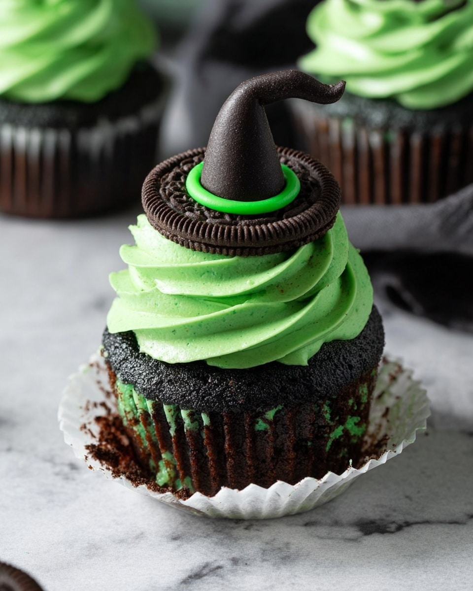 A dark black cupcake sits unwrapped on a white marbled surface, showing a moist, textured cake base with small chocolate pieces inside. It has one thick layer of bright green frosting on top, swirled smoothly, and a dark chocolate cookie placed flat on the frosting. On top of the cookie is a pointed black decoration shaped like a witch hat with a green frosting band at its base. In the background, there are more cupcakes with similar green frosting. Photo taken with an iphone --ar 4:5 --v 7
