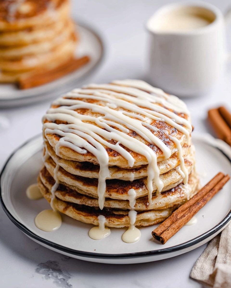 A stack of six thick pancakes sits on a white plate with a thin dark rim on a white marbled surface; the top pancake is swirled with a darker brown cinnamon-like pattern and drizzled with cream-colored icing in diagonal lines. A slice of the stack is cut away to show each pancake layer with light golden brown edges and a soft, fluffy inside with cinnamon swirls inside. The slice rests on a fork that is placed on the plate's edge. In the background, there is another similar stack blurred and a small white jug with cream or sauce. Photo taken with an iphone --ar 4:5 --v 7