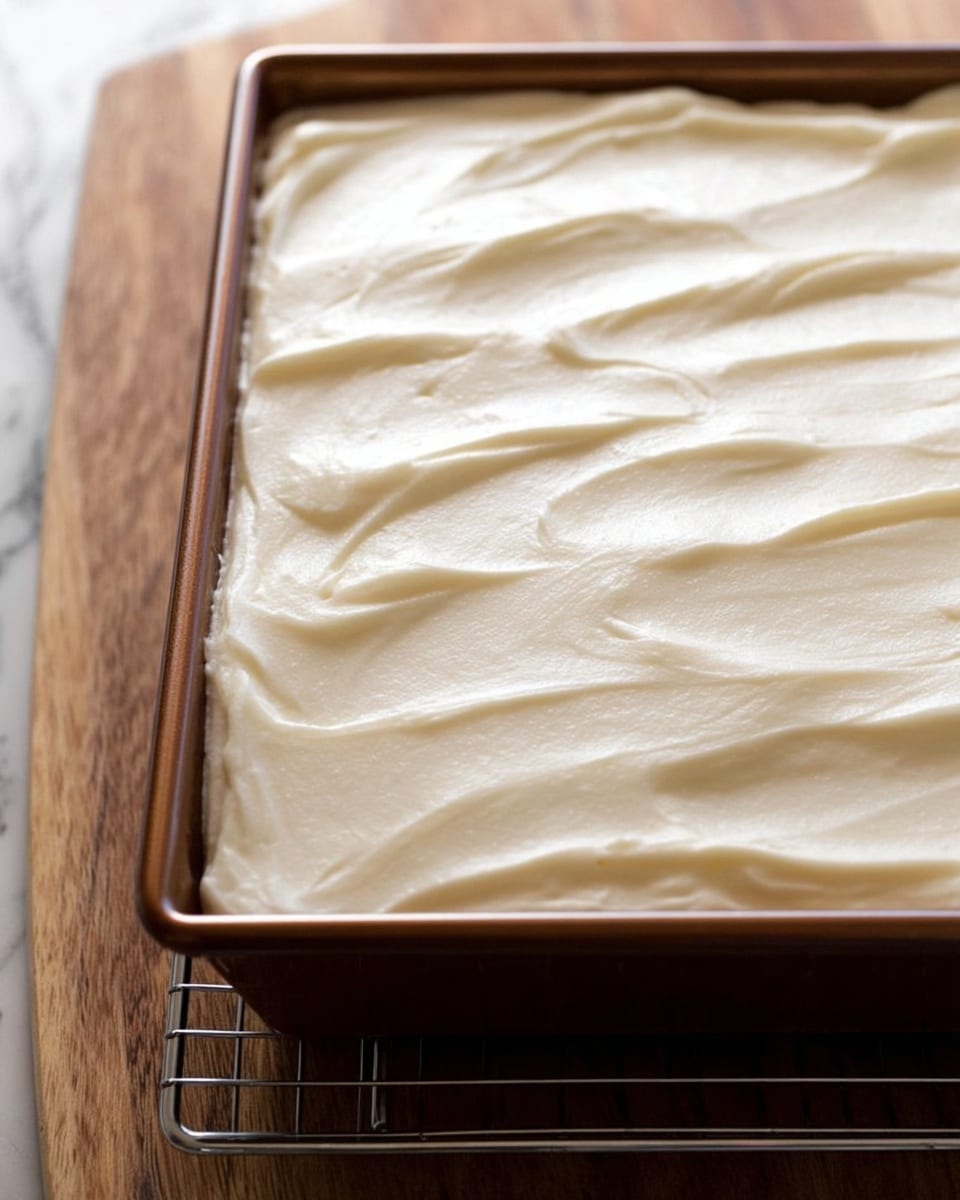 A single square slice of carrot cake sits on a white plate, showing two layers: the bottom layer is a moist, light brown carrot cake with small bits of carrot and darker spots, and the top layer is a thick, smooth, creamy white frosting spread evenly over the cake. Next to the cake on the plate is a silver fork with four tines. In the background, a white baking dish on a wire rack holds the rest of the carrot cake topped with the same white frosting, with one piece missing. The scene is set on a light wood table with a green cloth napkin that has a white and blue floral pattern, and the surface behind is a white marbled texture. Photo taken with an iphone --ar 4:5 --v 7