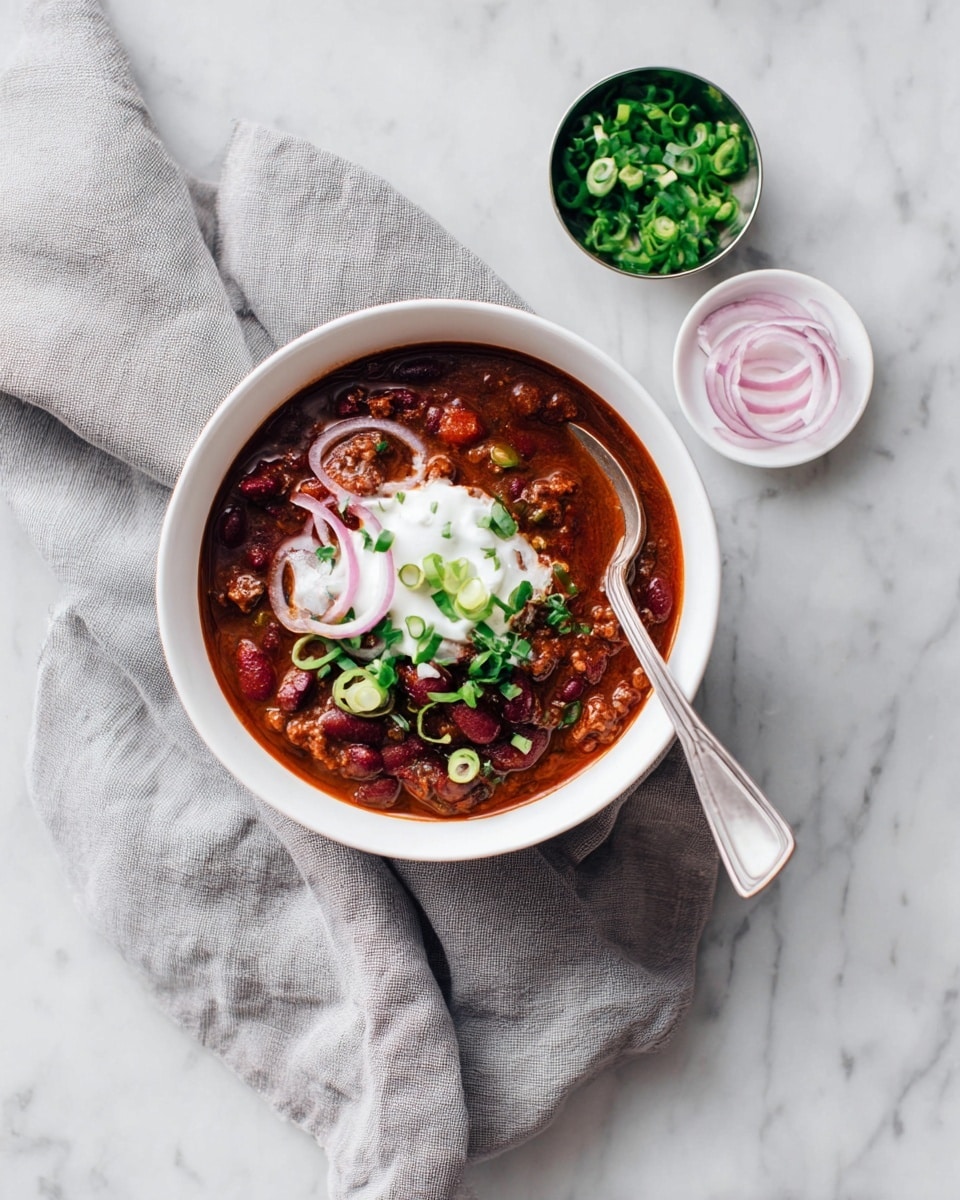 A white bowl filled with a thick chili stew showing dark red kidney beans and ground meat mixed in a rich red sauce as the first layer, topped with a smooth white dollop of sour cream as the second layer, scattered with bright green chopped spring onions and some thin pale pink onion rings as the third layer, a silver spoon resting inside the bowl, placed on a wrinkled gray cloth on a white marbled surface. Nearby, a small white dish holds additional pale pink onion rings, and a small round container is filled with more bright green chopped spring onions. Photo taken with an iphone --ar 4:5 --v 7