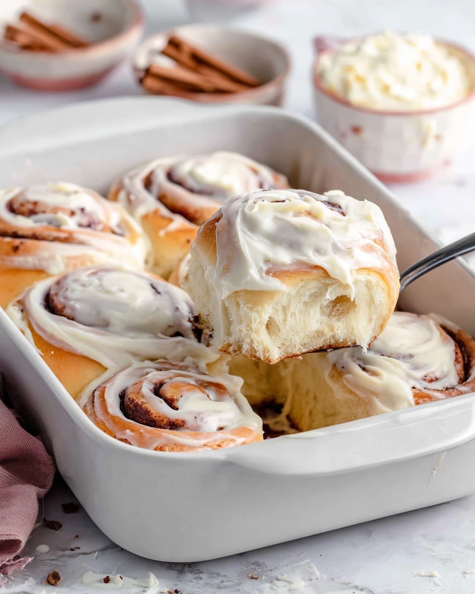The image shows a white rectangular baking dish filled with seven cinnamon rolls, each topped with thick, creamy white icing. The rolls have a soft, fluffy texture with golden brown edges and visible swirls of cinnamon inside. One cinnamon roll is being lifted by a silver utensil, revealing the soft inside and the white icing melting slightly over the sides. The baking dish rests on a white marbled surface, and the background is softly blurred, focusing attention on the warm, inviting rolls. photo taken with an iphone --ar 4:5 --v 7