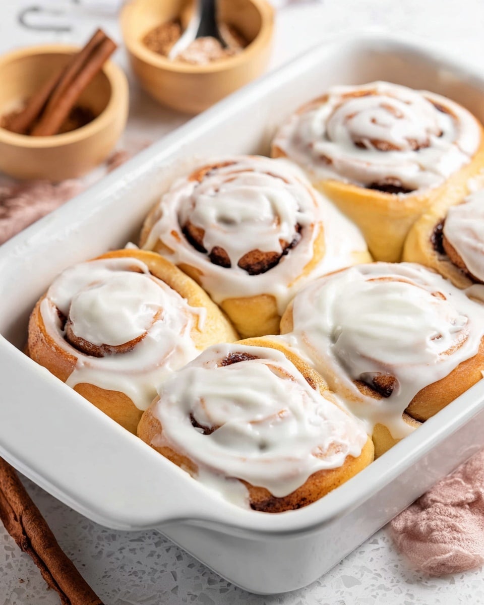 The image shows a single cinnamon roll placed on a white plate set on a white marbled surface. The cinnamon roll has two visible layers: the base layer is a golden-brown soft dough with fluffy texture and the top layer is thick white cream cheese frosting generously spread, slightly melting over the dough edges. The roll itself has a swirl pattern where the dough curls around cinnamon filling, creating an orange-brown spiral inside the white frosting. In the blurred background, there is another white plate with a similar cinnamon roll and part of a white baking dish filled with more cinnamon rolls, slightly out of focus. Photo taken with an iphone --ar 4:5 --v 7