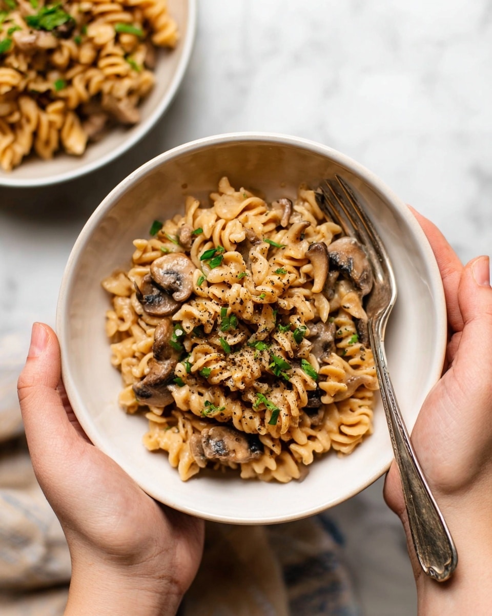 A close-up view of a pot filled with creamy rotini pasta mixed with sliced mushrooms, all coated in a thick brown sauce. The pasta spirals are golden beige with a glossy texture, while the mushrooms are soft with light brown tops and pale undersides, scattered evenly throughout the dish. A black spoon with a light wooden handle is partially submerged in the pasta, stirring it gently. The pot is light beige with small speckles inside, resting on a white marbled surface. Small bits of black pepper are sprinkled on top, adding texture to the creamy mixture. Photo taken with an iphone --ar 4:5 --v 7