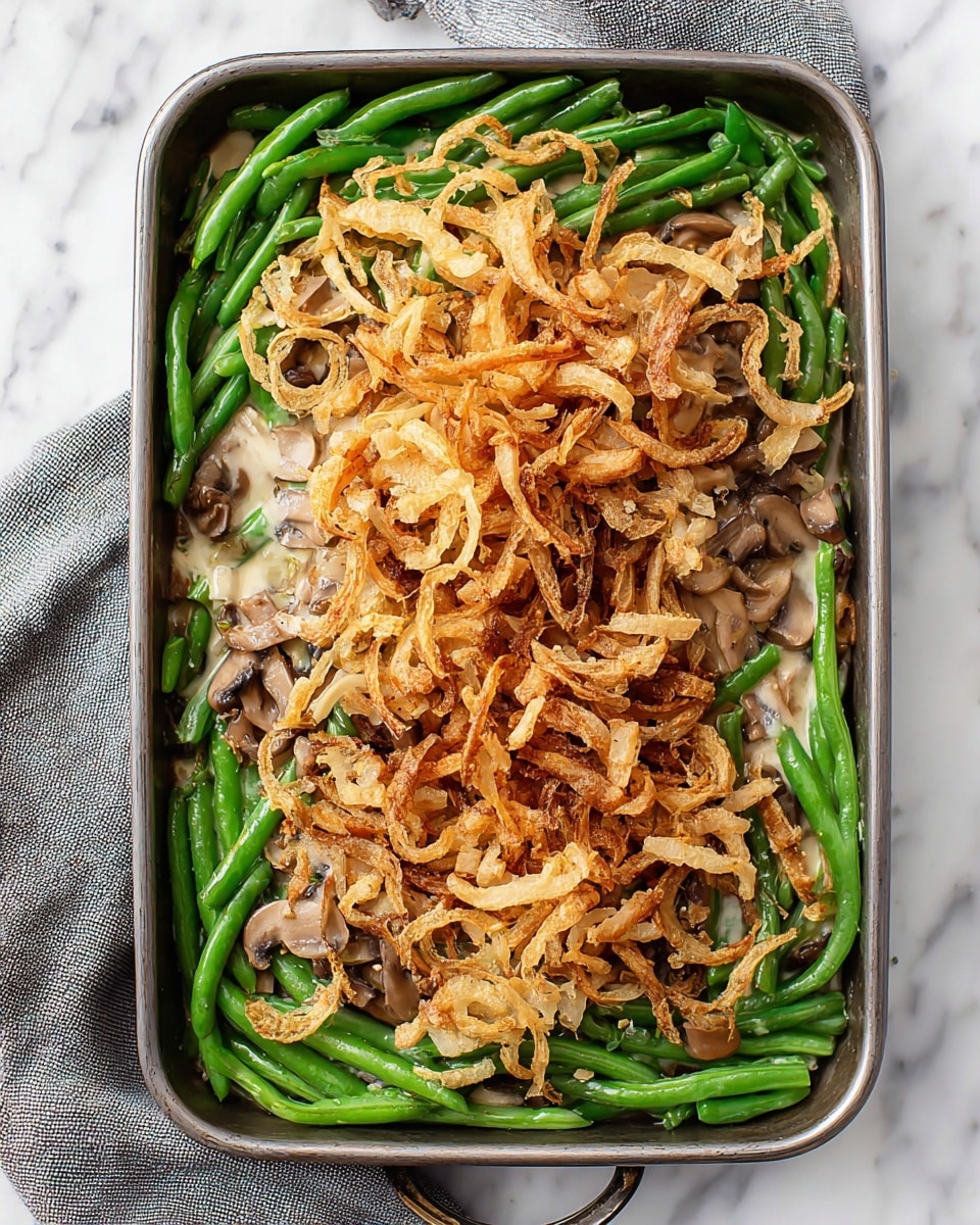 A rectangular metal baking dish holds a green bean casserole with three distinct layers: the bottom layer is bright green cooked green beans, the middle layer is a creamy light beige mushroom sauce with visible small mushroom slices scattered throughout, and the top layer consists of golden-brown crispy fried onion strips piled thickly, some slightly curled with a crunchy texture. The dish is set on a white marbled surface with a gray and white cloth nearby. Photo taken with an iphone --ar 4:5 --v 7