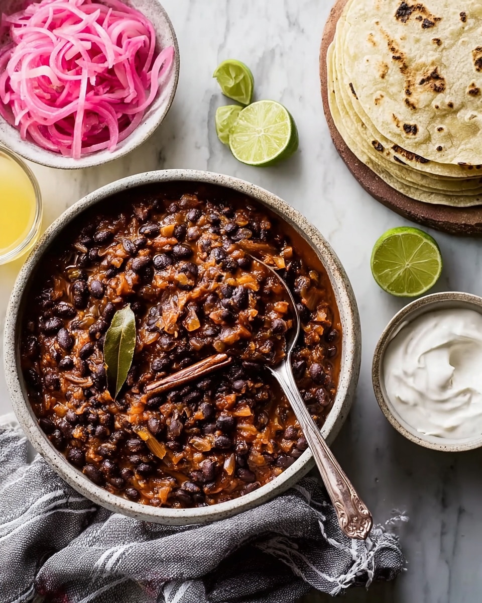 The image shows a round white bowl filled with a dark reddish-brown bean stew that includes black beans, diced vegetables, and visible bay leaves on top, with a spoon resting inside the bowl. To the top right is a white bowl holding bright pink pickled onions with thin slices visible. Nearby, to the right, there are three folded tortillas stacked on a wooden board, accompanied by lime wedges placed around them. A small white bowl with smooth white sour cream and a spoon is on the lower right edge. Two clear glasses filled with a pale yellow liquid are visible at the top left and bottom right of the image. The entire scene is set on a white marbled surface, and a beige and brown textured cloth is placed under the bowl of the bean stew. photo taken with an iphone --ar 4:5 --v 7