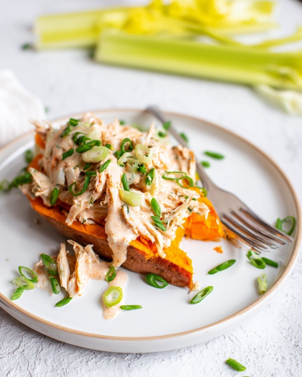 The dish shows one slice of orange roasted sweet potato placed on a white plate with a yellow and white striped cloth underneath, all on a white marbled surface. On top of the sweet potato is a layer of light shredded meat mixed with a bright orange sauce, drizzled with small drops of white creamy sauce. The dish is garnished with green chopped chives scattered around and on top. Next to the plate, there are two stalks of pale yellow celery. A silver fork rests to the right side of the plate. In the background, a blurred white plate with a similar sweet potato dish is visible. Photo taken with an iphone --ar 4:5 --v 7