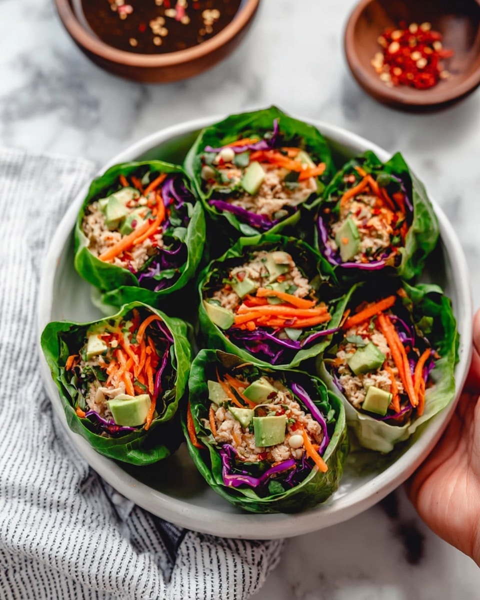 This image shows six fresh lettuce wraps arranged in a white bowl. Each wrap has three clear layers: a dark green leafy outer layer, a vibrant purple shredded cabbage layer next, and an inner mix of bright orange shredded carrots, light green avocado slices, and beige cooked filling at the center. The bowl sits on a white marbled surface with a striped cloth nearby. A small wooden bowl with dark sauce and red pepper flakes is placed next to the bowl of wraps. photo taken with an iphone --ar 4:5 --v 7