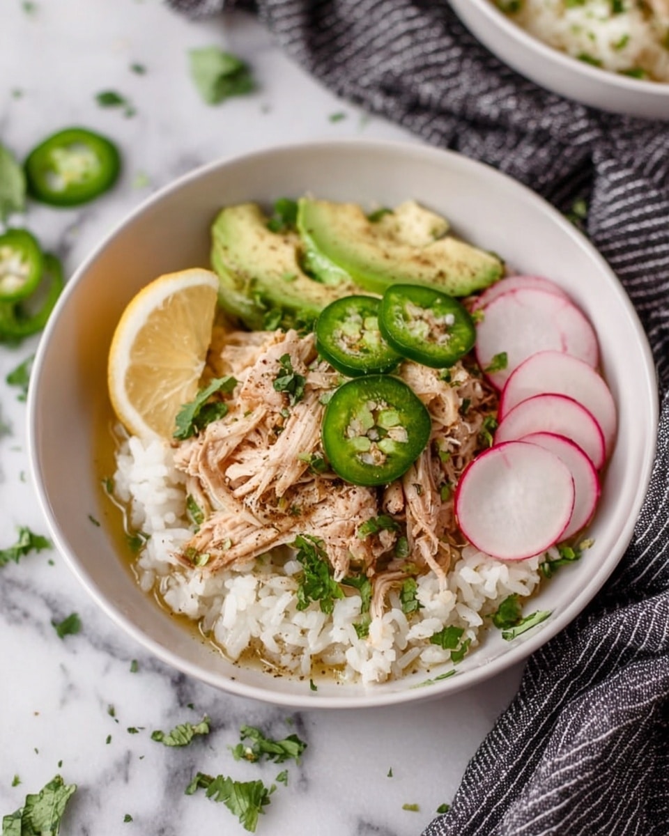 A shallow white bowl holds the dish, set on a white marbled surface with a black and white striped cloth nearby. The base layer is soft white rice spread evenly across the bowl. On top, there is a layer of shredded light tan chicken scattered in the center. A few slices of bright green jalapeño peppers and pale green avocado are placed over the chicken. Bright pink and white radish slices are on the right side, and a wedge of pale yellow lemon rests on the left. Small green cilantro leaves are sprinkled around for color. The image is softly lit, showcasing the fresh ingredients clearly, photo taken with an iphone --ar 4:5 --v 7