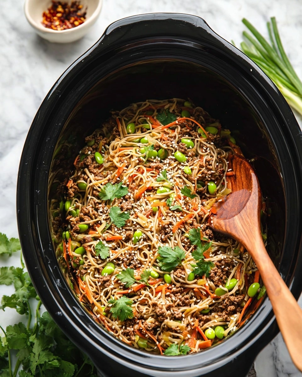 The image shows a black slow cooker filled with thin white noodles mixed with light brown ground meat, thin orange carrot strips, green edamame beans, and sliced green onions. There are also some sesame seeds and fresh cilantro leaves scattered on top. A wooden spoon rests inside the slow cooker, partially buried in the noodle mix. Around the cooker, a white marbled surface is visible with small bowls holding spices and fresh herbs nearby. Photo taken with an iphone --ar 4:5 --v 7