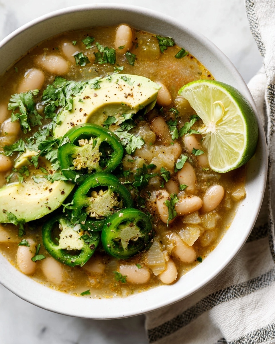A white plate holds a layered dish starting with a base of soft beans and cooked vegetables in a light broth. On top of this, there are fresh slices of green avocado placed in small chunks. Next, bright green jalapeño rings with visible seeds are spread around. Fresh cilantro leaves add a darker green color, giving a fresh look. A wedge of lime with a sprinkle of black pepper sits on one side. The plate is set on a white marbled surface with a striped cloth nearby. photo taken with an iphone --ar 4:5 --v 7