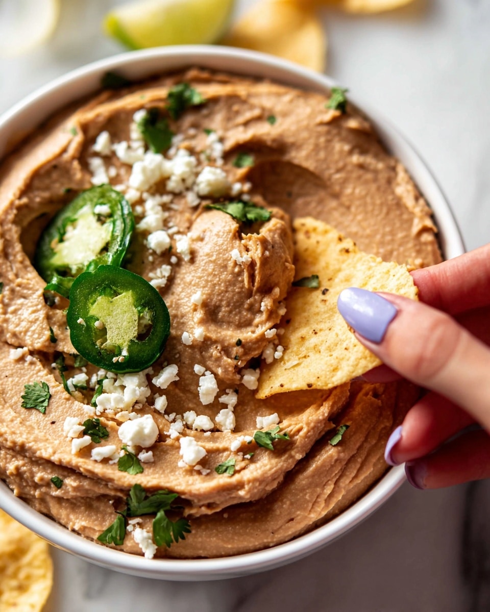 A close-up of a thick, creamy brown spread with a smooth, slightly swirled texture, spread out evenly in a white shallow bowl. On top, there are small white crumbly cheese pieces and bright green cilantro leaves scattered around, along with a thin round slice of green jalapeño with visible seeds. A woman's hand with light purple nail polish is dipping a pale yellow, lightly toasted tortilla chip with some brown spots into the spread. The bowl is placed on a white marbled surface. photo taken with an iphone --ar 4:5 --v 7