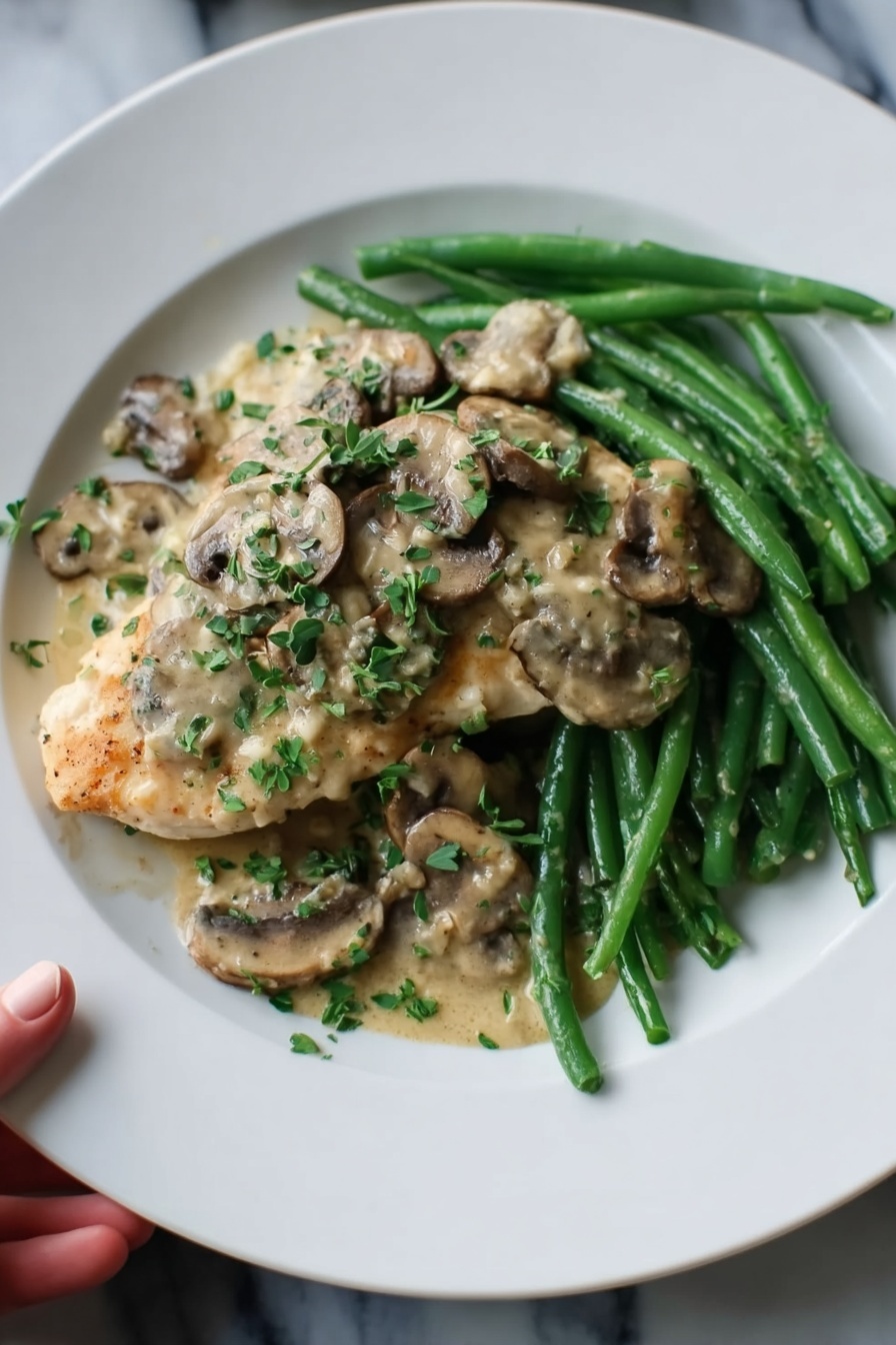 The dish shows a white plate with a bed of green beans at the back, bright and crisp, arranged in a neat pile. On top of the green beans, there is a piece of light golden cooked chicken, covered generously with creamy beige mushroom sauce. The sauce includes sliced mushrooms that look soft and well cooked, scattered over the chicken and green beans. Finely chopped fresh green herbs are sprinkled over the dish giving a fresh touch. The plate sits on a white marbled surface, and a woman's hand is holding the edge of the plate from the left side. photo taken with an iphone --ar 2:3 --v 7 - Easy Chicken Marsala Without Wine, Chicken Marsala Without Wine, Alcohol-Free Chicken Marsala, Quick Chicken Marsala Dinner, Healthy Chicken Marsala Recipe