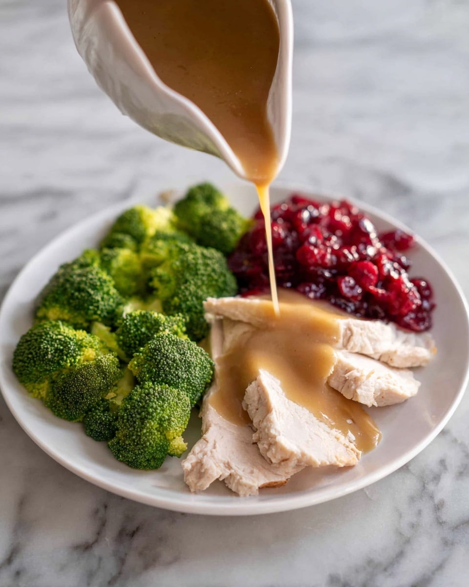The image shows a white plate with four main layers. At the bottom, there are slices of cooked turkey, pale beige and soft in texture, spread across the center of the plate. On top of the turkey is a thick layer of light brown gravy with mushroom pieces, covering most of the meat. To the left side of the plate, there are several bright green broccoli florets with a fresh, bumpy texture. On the right side, there is a small serving of dark red cranberry sauce with a slightly chunky texture. A small sprig of fresh green parsley is placed near the broccoli for garnish. The plate is set on a white marbled surface. Photo taken with an iphone --ar 4:5 --v 7