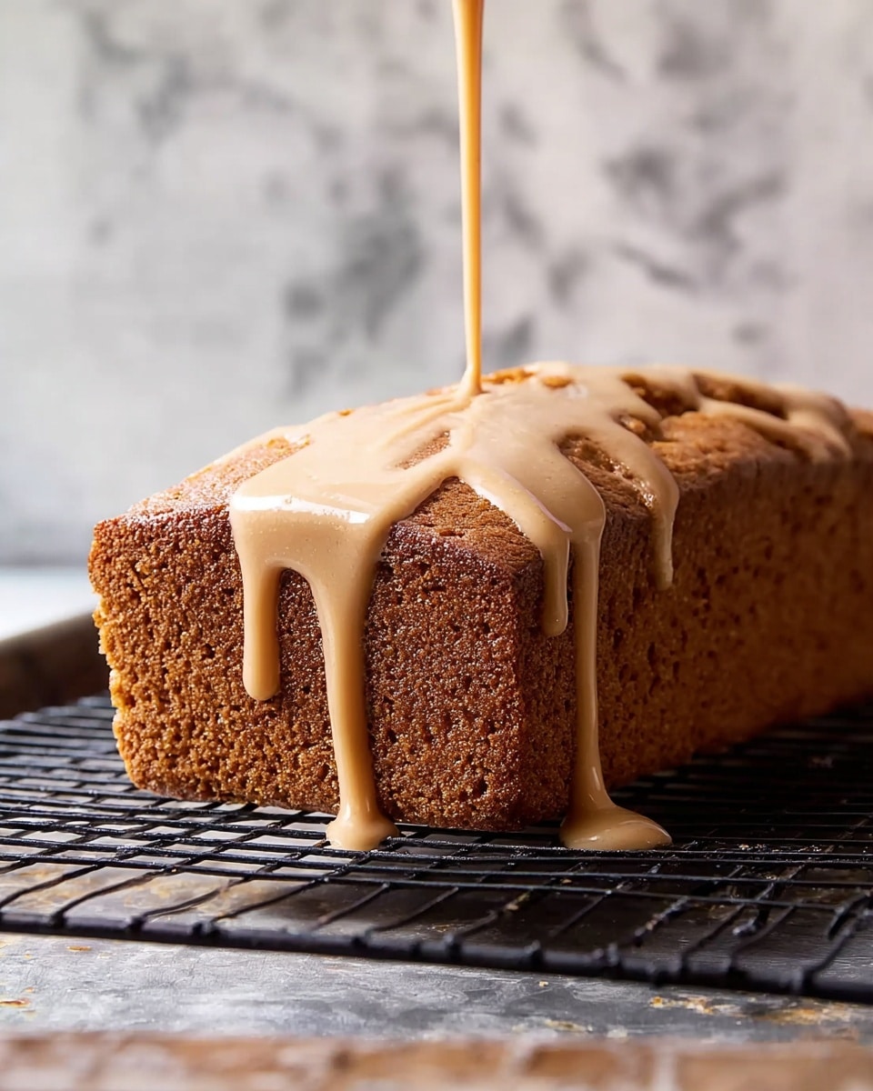 A single rectangular brown cake loaf with a slightly rough and textured surface sits on a black metal cooling rack. A light tan creamy glaze is being poured over the top center of the cake, flowing down the sides with a smooth and glossy texture, creating thick drips hanging off the edges. The background shows a white marbled surface slightly blurred for focus on the cake and glaze. Photo taken with an iphone --ar 4:5 --v 7