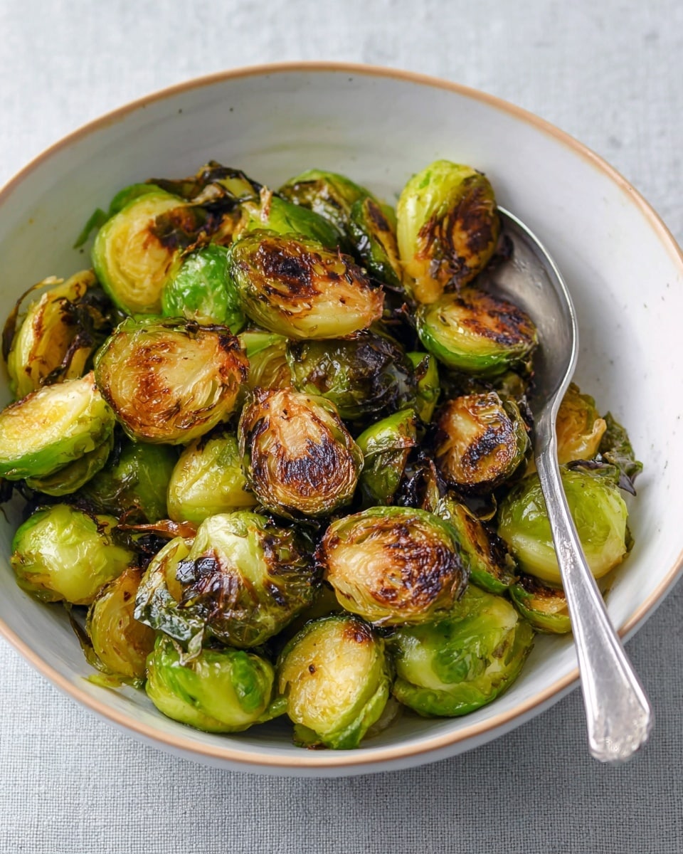 A white bowl filled with roasted Brussels sprouts, showing two layers: the bottom layer is bright green, halved Brussels sprouts with a soft texture, while the top layer is made of dark brown, crispy Brussels sprout leaves scattered unevenly on top. There is a silver spoon resting inside the bowl on the right side. The bowl sits on a cloth with a soft blue-gray color, and the surface under the cloth has a white marbled texture. Photo taken with an iphone --ar 4:5 --v 7