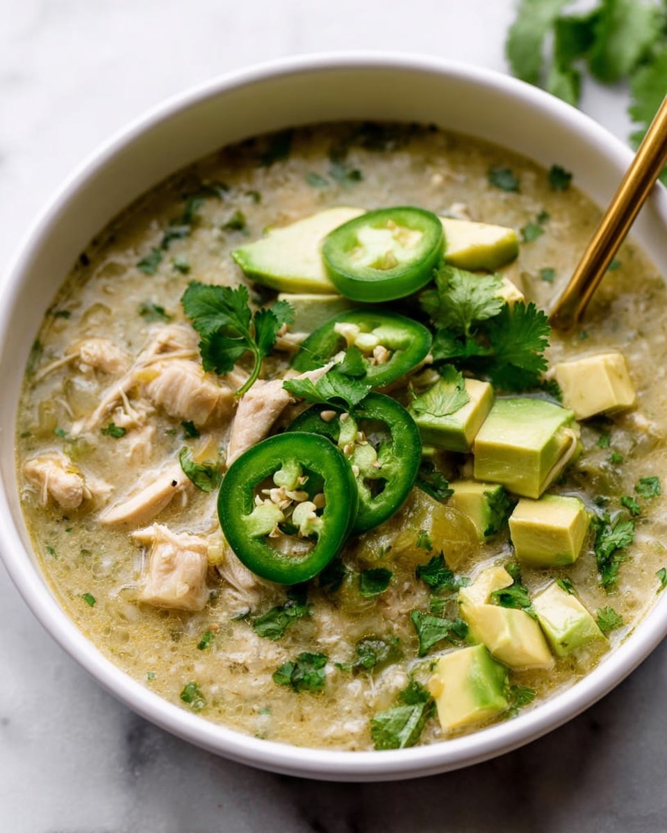 A white bowl filled with a thick greenish broth containing chunks of light beige cooked chicken and bits of herbs mixed in. On top, there are three bright green slices of jalapeño pepper placed in the center, next to thin slices of avocado showing a pale yellow and light green gradient. Fresh coriander leaves with their jagged edges are scattered mostly on one side of the bowl. A golden spoon is placed partly inside the bowl, resting near the chicken pieces. The bowl sits on a white marbled surface with a soft background. Photo taken with an iphone --ar 4:5 --v 7