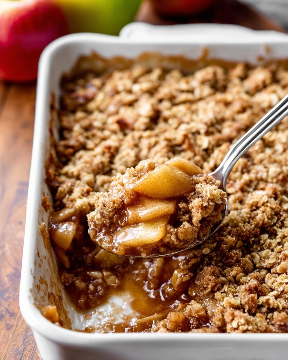 The image shows a white rectangular baking dish filled with apple crumble. The dessert has two clear layers: the bottom layer is made of soft, cooked apple pieces in a shiny, golden-brown syrup, while the top layer is a thick, crumbly oat topping with a mix of light and dark brown shades and a coarse texture. A silver spoon is scooping some apple crumble from the right side, revealing the apple filling underneath. The dish sits on a dark wooden surface with a slight textured look. Photo taken with an iphone --ar 4:5 --v 7
