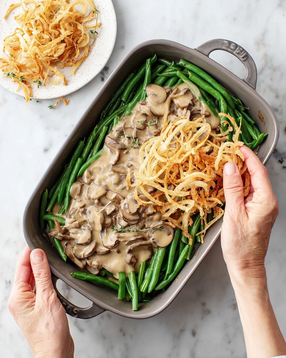 A black rectangular baking dish filled with three layers: the bottom layer is bright green fresh green beans arranged neatly around the edges; the middle layer is a creamy beige mushroom sauce spread over the green beans with visible sliced mushrooms; the top layer consists of golden crispy fried onions being placed by a woman's hands, which are holding a handful of the onions above the dish. Next to the baking dish, on the right, there is a small wooden plate with more crispy fried onions on a white marbled surface background. Photo taken with an iphone --ar 4:5 --v 7
