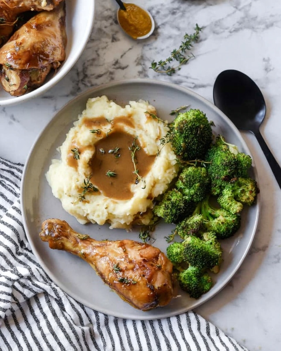 A white plate holds a simple meal with three main parts arranged in sections: mashed potatoes on the top left with a light brown gravy pool in the center, two brown baked chicken drumsticks with a slight green herb garnish placed below the mashed potatoes, and a cluster of bright green steamed broccoli on the right side. The plate sits on a white marbled surface with a striped white and gray cloth nearby and some thyme sprigs lying next to the plate. A small dollop of light yellow mustard and a spoon are visible at the top right corner. photo taken with an iphone --ar 4:5 --v 7
