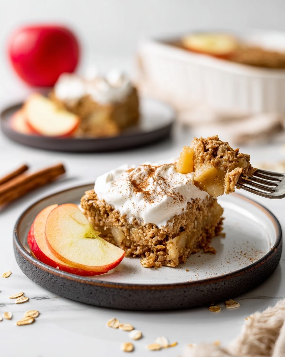 The image shows a single square piece of apple oatmeal cake on a round white plate with a black rim. The cake has two clear layers: the bottom layer is light beige and soft, while the top layer is chunky with visible oats and small pieces of cooked apple mixed in, sprinkled with cinnamon. A generous dollop of white whipped cream is on top of the cake, lightly dusted with cinnamon. Next to the cake on the plate are two thin apple slices with red skin and pale yellow flesh. A silver fork rests on the plate with a forkful of cake, showing the crumbly texture. In the background, there is a blurred whole red apple, a white bowl with more yellow apple slices, and another plate holding a similar piece of cake. The surface is a white marbled texture with scattered oats and cereal crumbs. photo taken with an iphone --ar 4:5 --v 7
