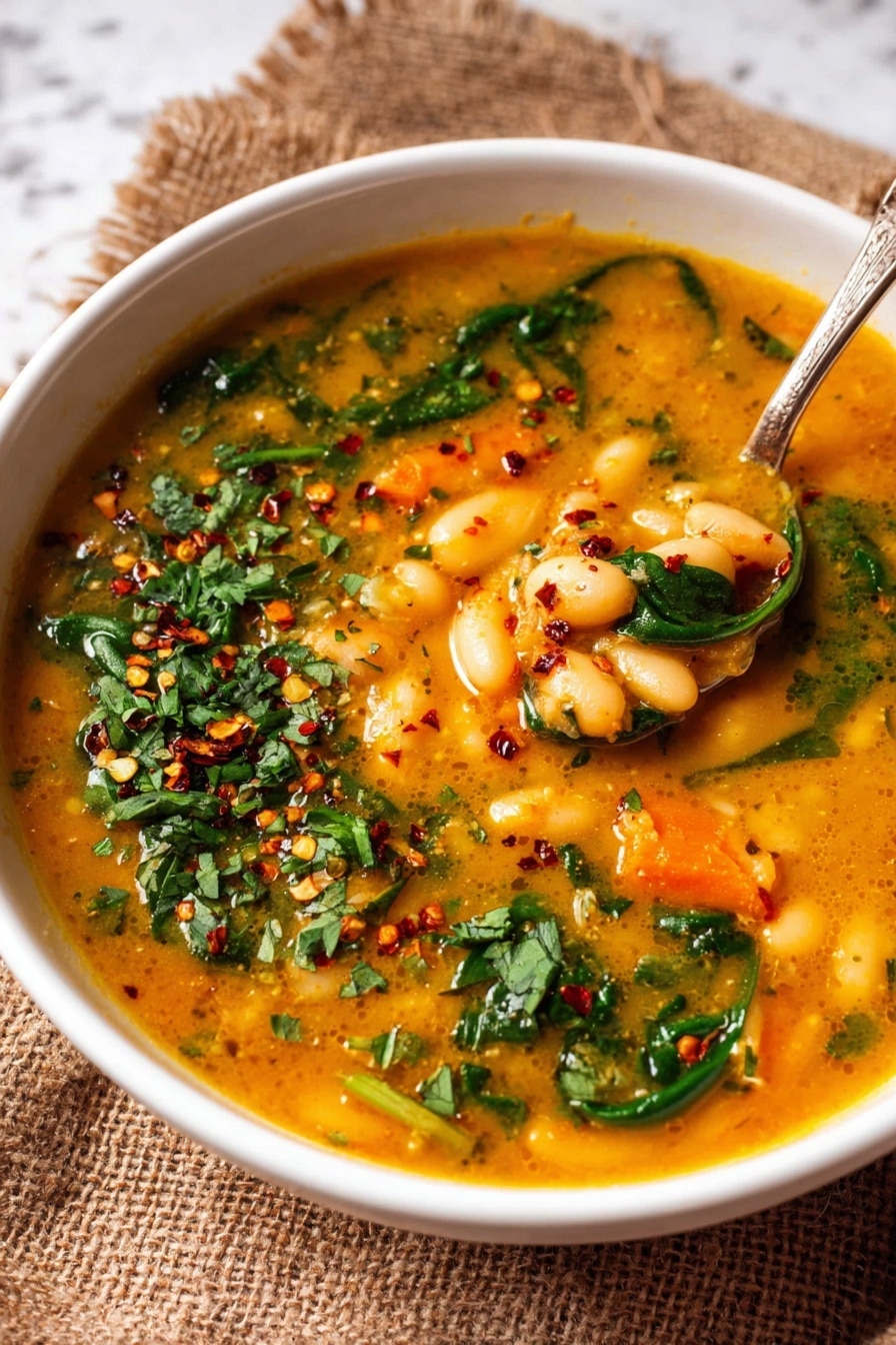 A white bowl filled with a thick orange-yellow soup that has white beans and small pieces of orange carrots visible throughout. There are bright green spinach leaves and chopped green herbs spread on top and inside the soup. A spoon resting in the bowl holds some of the bean and carrot mixture. The soup surface is sprinkled with red chili flakes and green herbs, adding color contrast. The bowl sits on a textured brown fabric over a white marbled surface. Photo taken with an iphone --ar 2:3 --v 7 - Hearty Tuscan White Bean Soup, white bean soup, Tuscan bean soup recipe, hearty Italian bean soup, quick veggie soup