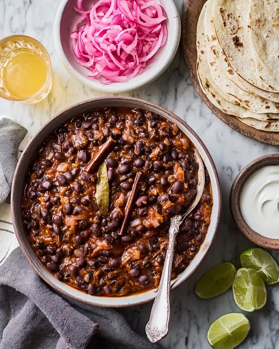 A white bowl filled with a thick black bean stew featuring dark black beans mixed with small pieces of orange and green vegetables and two bay leaves on top, a spoon resting inside the bowl. Nearby, a white bowl contains bright pink pickled onions, while folded white tortillas with dark spots are stacked on a wooden board beside lime wedges. A small white bowl of smooth white sour cream with a spoon sits on a white marbled surface along with glasses of light yellow olive oil, all arranged on a white marbled countertop. A striped cloth napkin is under the stew bowl, photo taken with an iphone --ar 4:5 --v 7