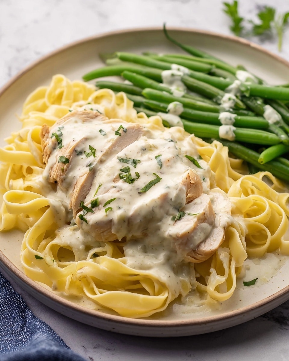 A white plate holds three main layers: the bottom layer is light yellow fettuccine pasta with a smooth texture, placed loosely across the plate. On top, there is a layer of sliced, pale chicken covered in a creamy white sauce with visible herbs. The chicken is arranged in slices, showing the sauce dripping slightly over the pasta. To the side, a small pile of vibrant green beans is also topped with a drizzle of the same creamy white sauce. Small green herb bits are sprinkled on top of the chicken and sauce. The plate is set on a white marbled surface. Photo taken with an iphone --ar 4:5 --v 7
